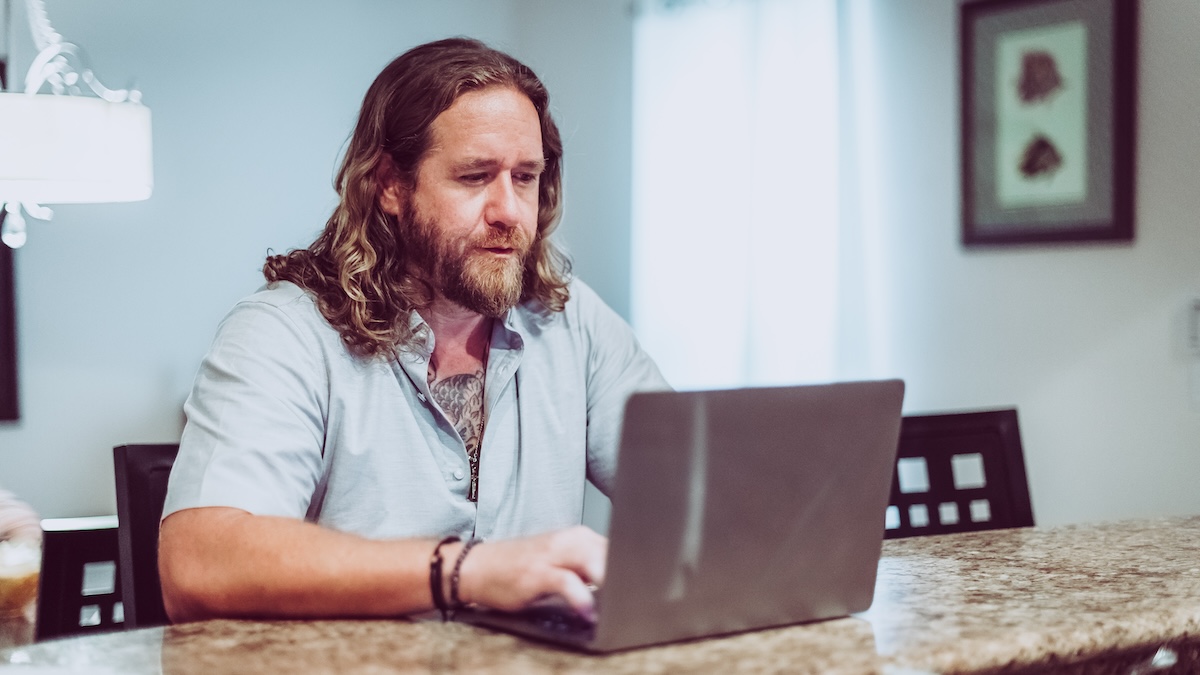 Man with beard and long hair works from home using a laptop at his kitchen counter. He is serious and concentrating on the screen, telecommuting