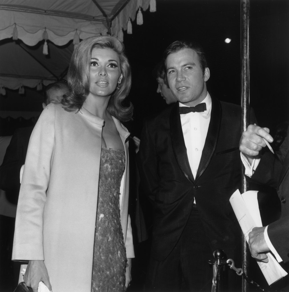 William Shatner, wearing a tuxedo, talks to his date, Nancy Kovack, while standing under a tent at a formal event.