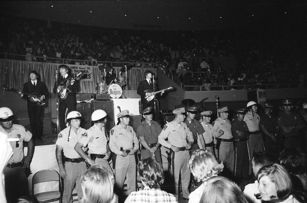 20th August 1964: A line of police officers prevent fans from getting too close, as The Beatles perform behind them on stage at the Convention Hall, Las Vegas.