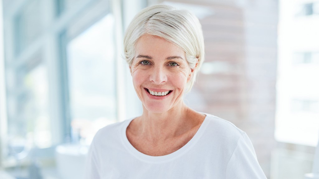 Gray-haired women who is standing in a bathroom and smiling. Her teeth look white after using a purple toothpaste