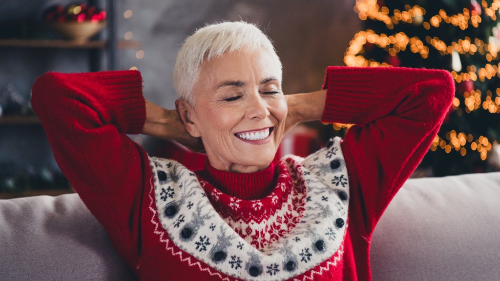 older woman relaxes on the sofa with Christmas tree in background; her eyes are closed as she smiles and savors this relaxing moment