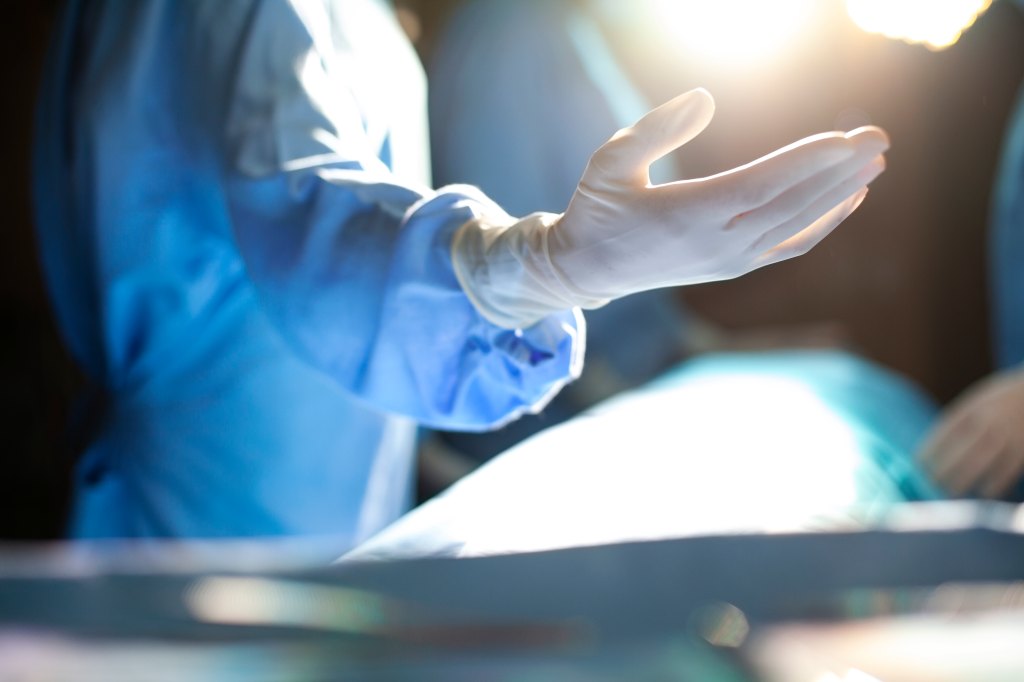 Hand of medical professional doctor surgeon wearing protective workwear latex glove reaching to colleague nurse in support care and teamwork in surgery operation room in hospital with colleagues and light