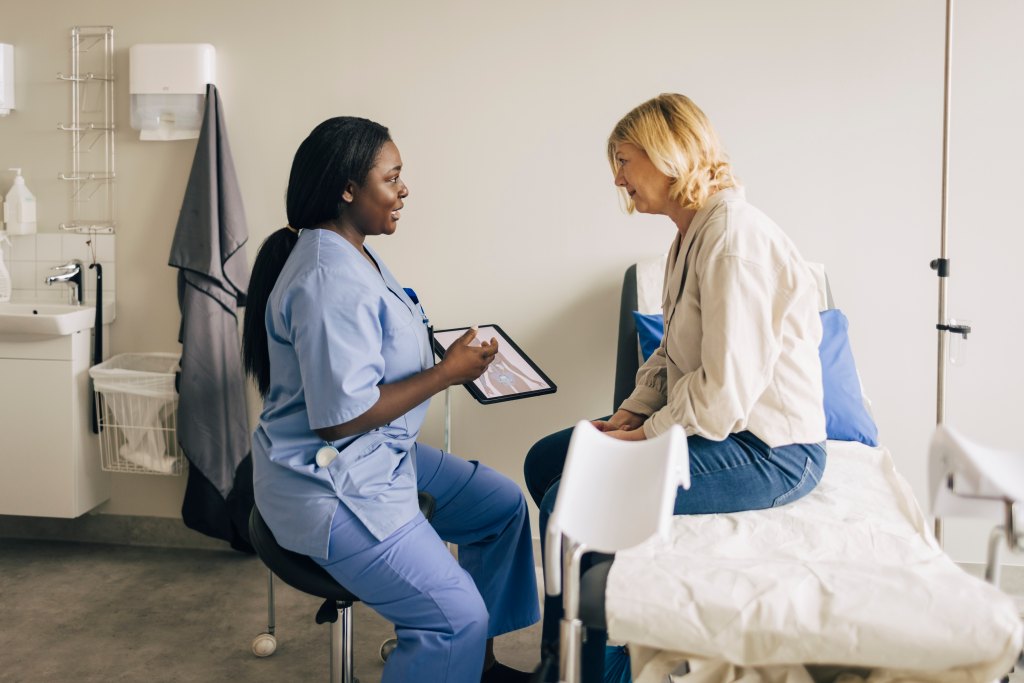 Gynecologist holding digital tablet and discussing with patient sitting on gurney in clinic