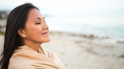 A woman with dark hair on a beach wrapped in a shawl with her eyes closed smiling