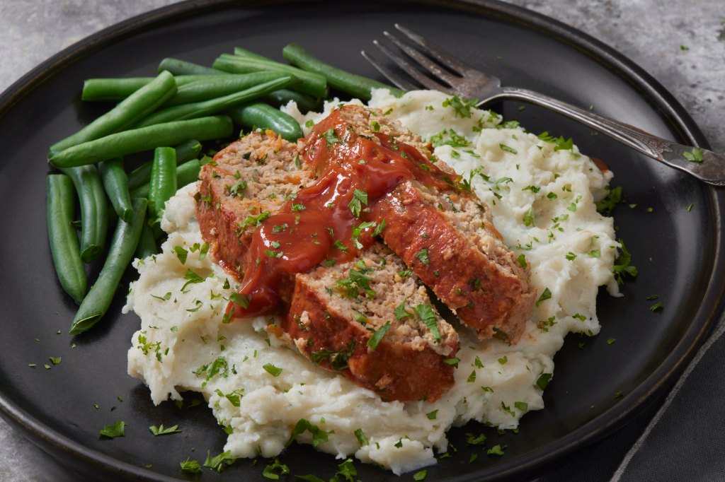 sliced meatloaf with ketchup sauce over mashed potatoes and green beans on plate