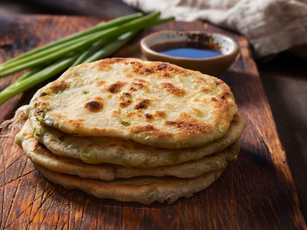 stack of flaky Chinese scallion pancakes for Lunar New Year with scallions and dipping sauce on cutting board