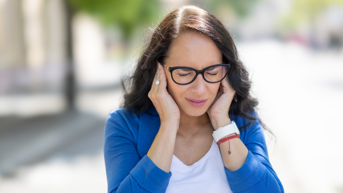 A woman with dark hair, a white shirt and blue cardigan closing her eyes and holding her hands to her ears to try to relieve sinus pressure