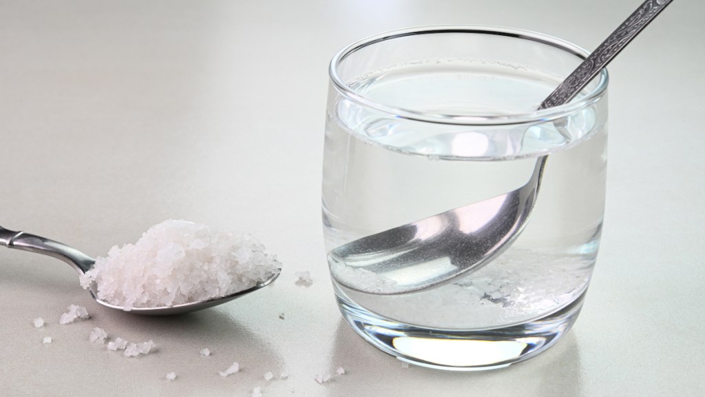 A glass of water with a spoon beside a spoon of salt, which helps stop bleeding gums