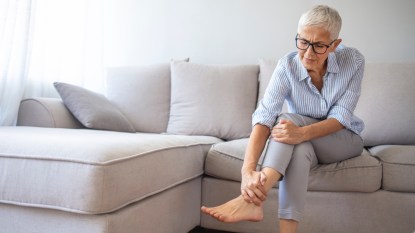 A woman with grey hair wearing a striped shirt sitting on a grey couch as she touches her painful cracked heels