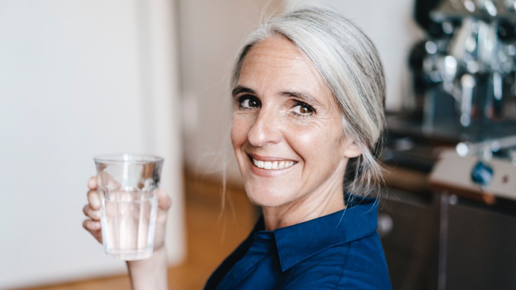 A woman with grey hear wearing a blue shirt holding a glass of water, which helps relieve sinus pressure in ears