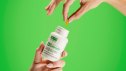 An image of natural appetite suppressant Calocurb being held by one hand and a pill plucked out of the bottle with another hand. The background of the image is light green.