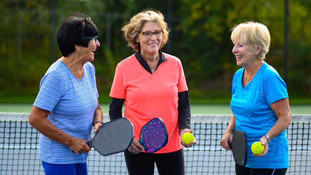 Three women on a court holding pickleball paddles and balls to fight stress belly