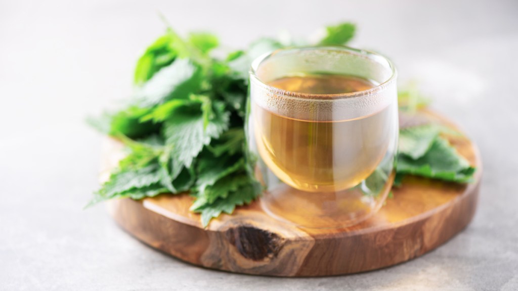 A cup of nettle tea on a wood board beside fresh nettle leaves