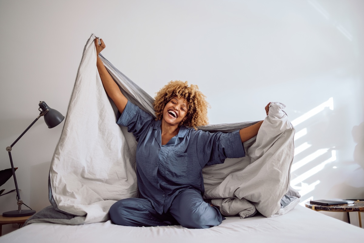Woman in her pajamas smiling and sitting on the bed while playing with bed covers. She is holding them on her back like a cape