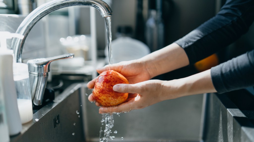 A close-up of a woman washing an apple under running water at the sink to avoid norovirus