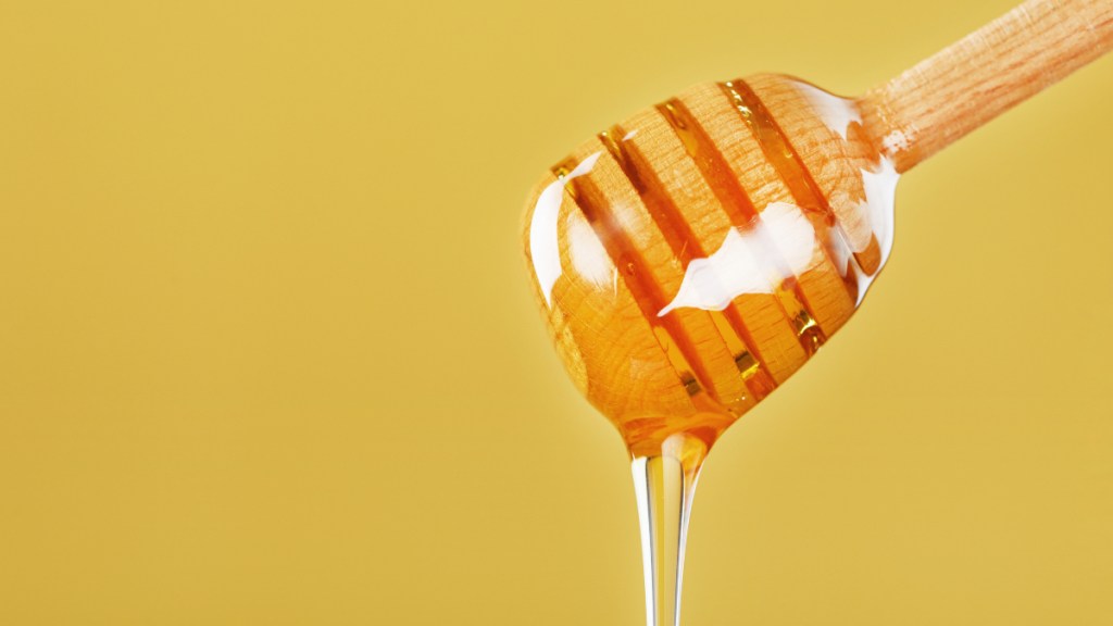 A wooden honey dipper with honey dripping off of it on a yellow background