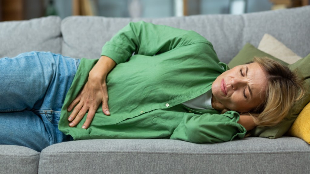 A woman in a green shirt lying on a grey couch holding her hand to her stomach in pain