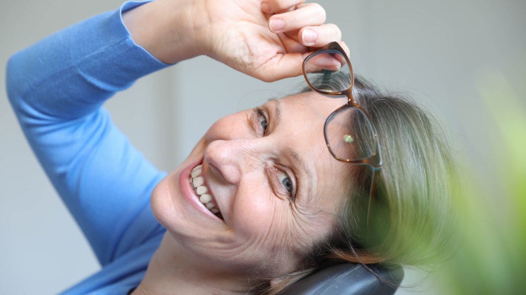 A woman in a blue shirt smiling and and leaning back while holding up her glasses to help get rid of a stye