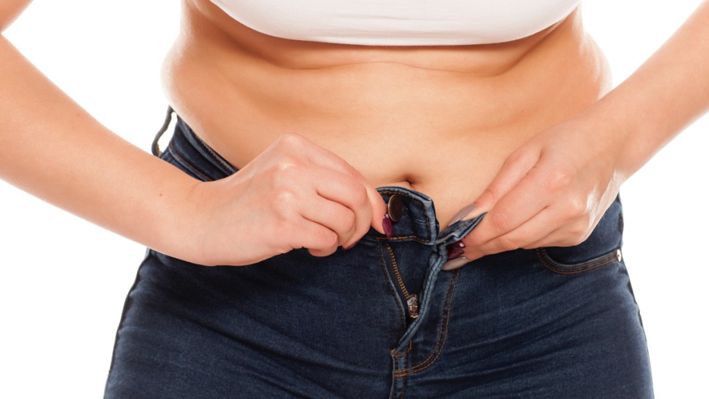 A close-up of a woman struggling to button her jeans due to stress belly