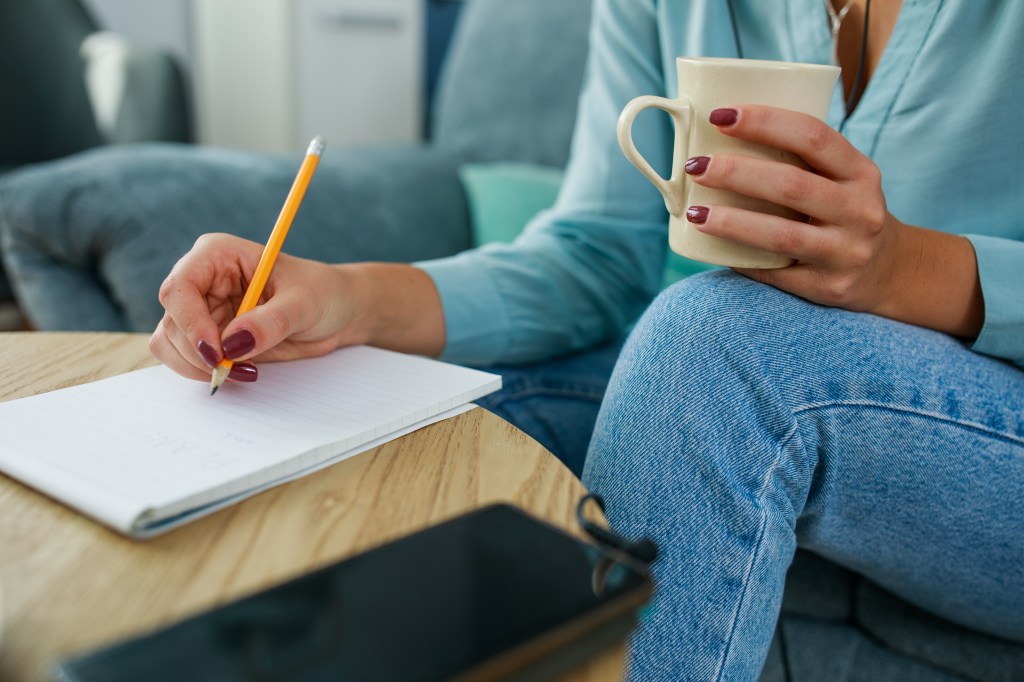 Woman writing in a notebook with right hand while holding coffee cup in left hand