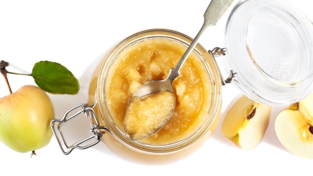 A jar of applesauce with a spoon beside fresh apples