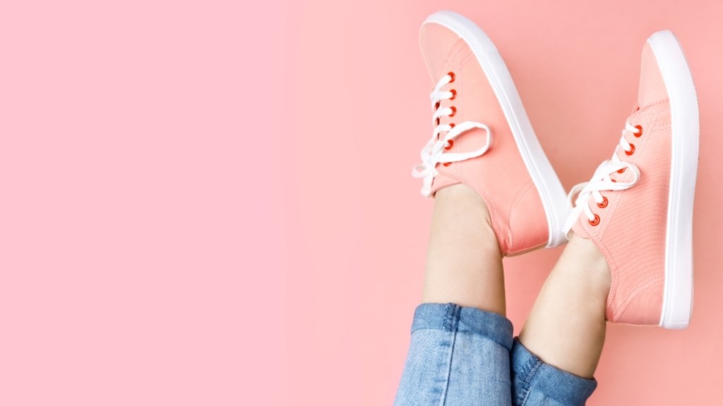 A close-up of a woman in jeans wearing pink sneakers against a pink background