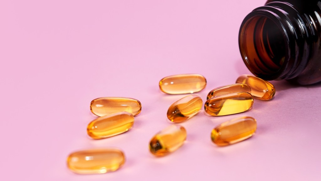 Omega 3 fish oil capsules coming out of an amber bottle against a pink background
