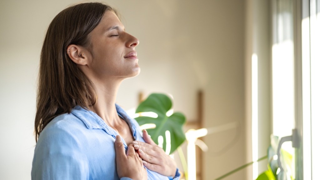 A woman in a blue shirt with brown hair closing her eyes while holding her hands to her chest to breathe deeply and block stress hives