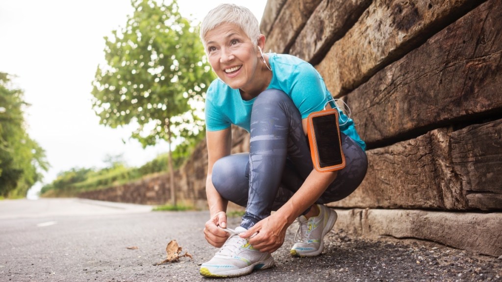 A mature, smiling woman in fitness attire lacing up her sneakers while outdoors 