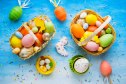 Close up photo of Easter baskets, colorful eggs, toy nest, cock doodle, hen and a mug on a blue table.