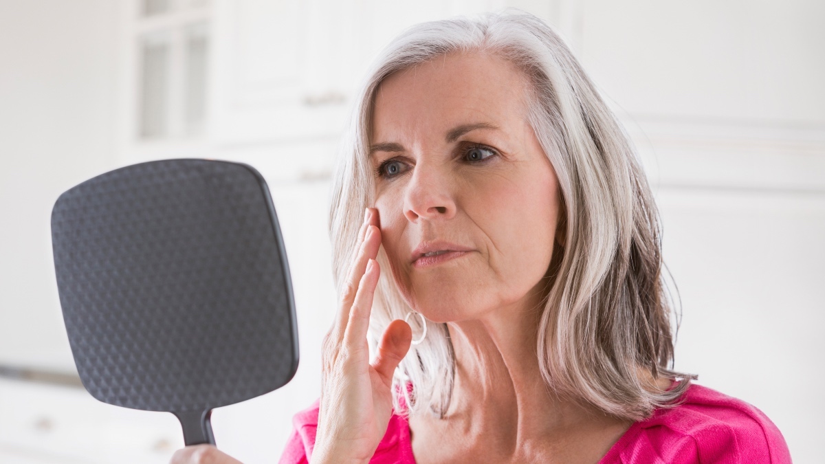 A mature woman wearing a pink shirt looking in a mirror while touching her hand to her face as part of her conjunctivitis self-care