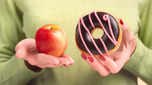 A close-up of a woman in a green shirt holding an apple in one hand and a donut in the other hand while using healthy eating hacks to decide what to eat