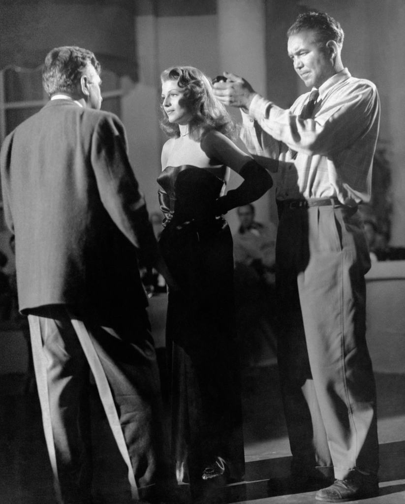 GILDA, director Charles Vidor, left, talks to Rita Hayworth, while a camera assistant checks the lighting, on-set, 1946