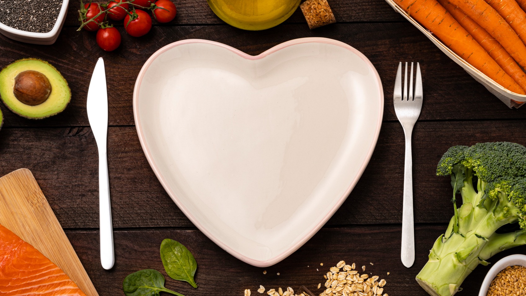 Top view of a heart shaped plate surrounded by a knife and a fork and some healthy foods for heart health