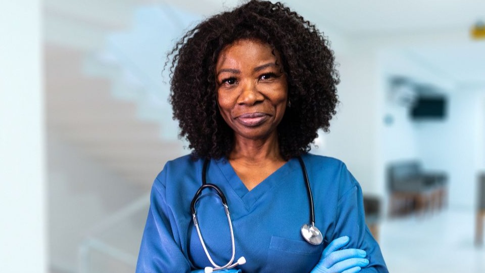 Smiling female nurse standing with arms crossed in hospital lobby wearing scrubs and gloves