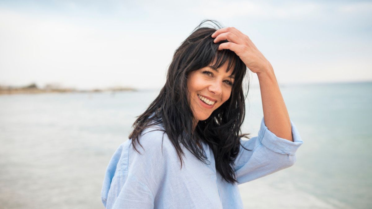 mature woman smiling while standing outdoors at a beach and holding her hand to her head