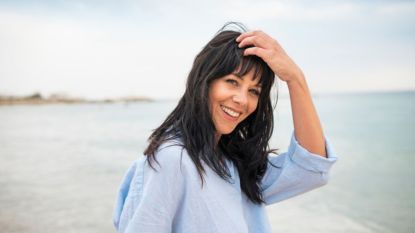mature woman smiling while standing outdoors at a beach and holding her hand to her head