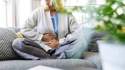 mature woman with stomach pain on a grey couch holding her abdomen due to stomach ulcer causes