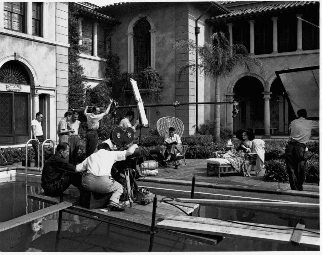 Gloria Swanson seated on set by a swimming pool during filming of Sunset Boulevard