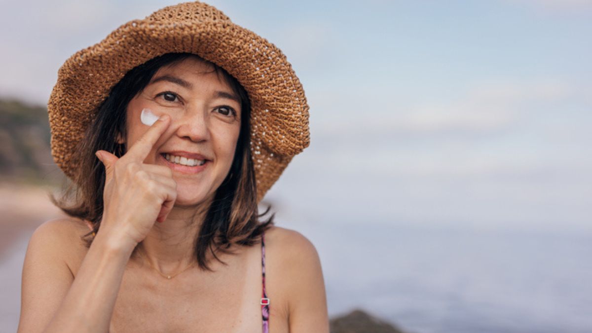 A mature woman wearing a hat and swimsuit while applying sunscreen to protect her psoriasis from summer sun damage