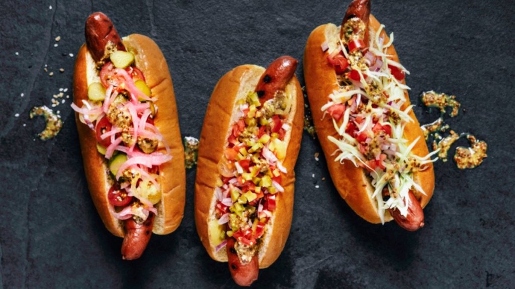 three smoked hot dogs with toppings in buns against black background