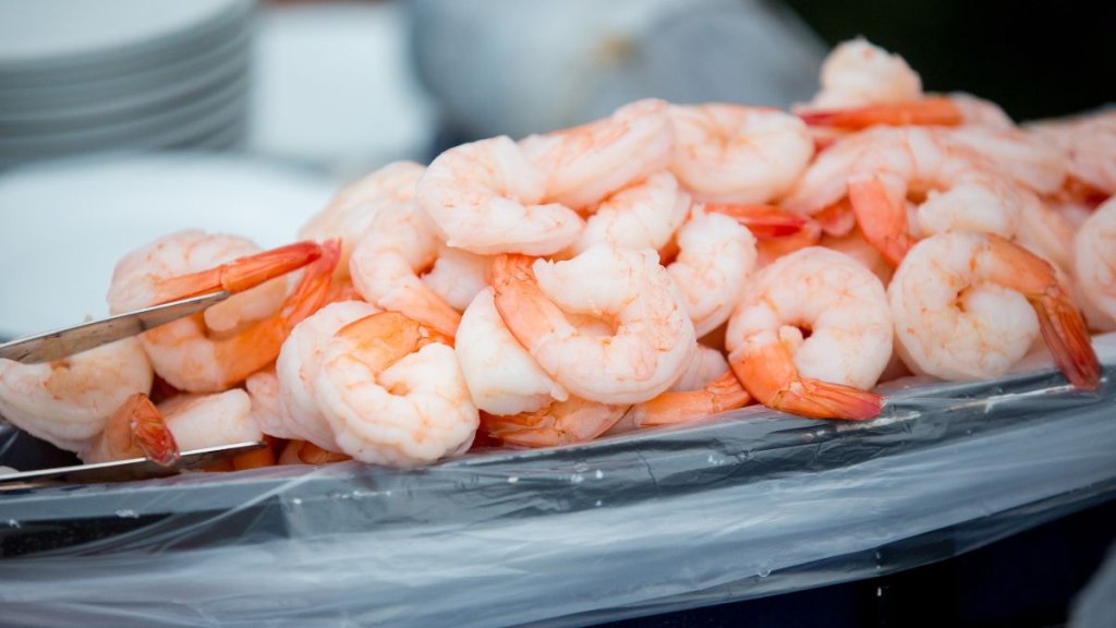 A heaping tray of peeled, cooked shrimp ready to be served on ice, captured at an outdoor seafood raw bar with white plates stacked in the background