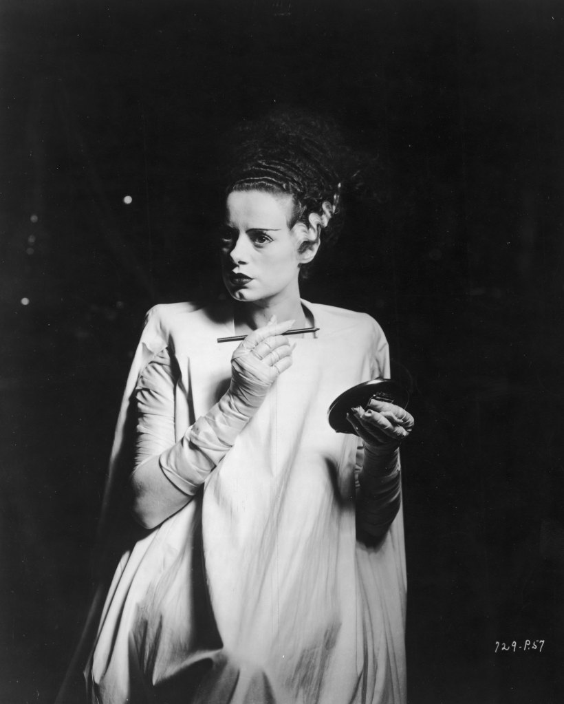 1935: British actor Elsa Lanchester (1902 - 1986) stands in a spotlight surrounded by darkness while holding an eyebrow pencil and a compact mirror on the set of director James Whale's film, 'The Bride of Frankenstein'. She wears a wig with a gray streak and a light-colored smock