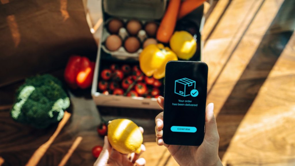 Female hands confirming the arrival of a box of home delivery filled with multi-coloured fresh and healthy organic groceries ordered online with mobile app on smartphone. Grocery subscription boxes. Meal kits. Convenient and time savings for daily life