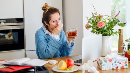 Portrait of woman with sniffles sitting indoors in kitchen, using laptop.