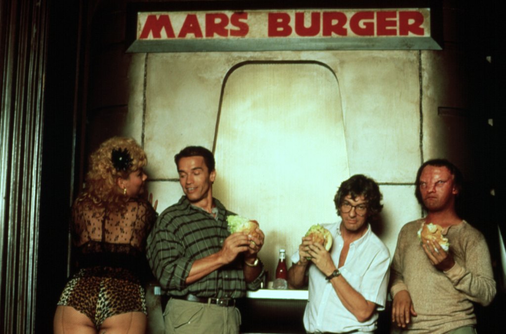 TOTAL RECALL, Arnold Schwarzenegger (left of center), Paul Verhoeven (right of center) feast on Mars burgers with extras, on set, 1990