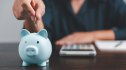 woman sits at desk with calculator and places a coin in a piggy bank