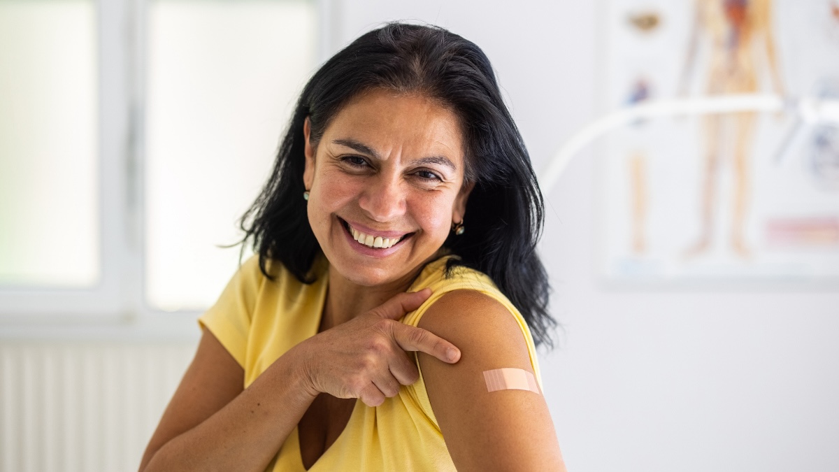 middle-aged woman smiles as she points to her upper arm, where a small adhesive bandage covers the area where she received her pneumonia vaccine