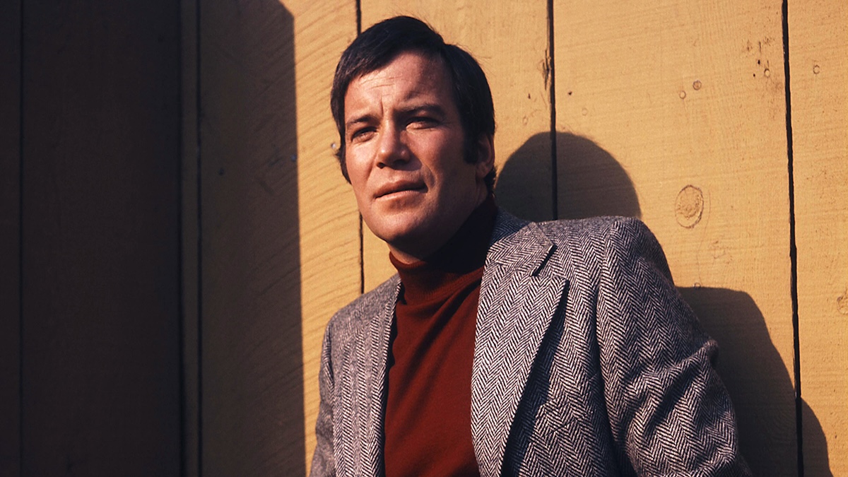 Canadian actor William Shatner, wearing a dark red turtleneck sweater beneath a grey herringbone tweed jacket, his arms folded as he stands against a wooden fence, United States, circa 1983.