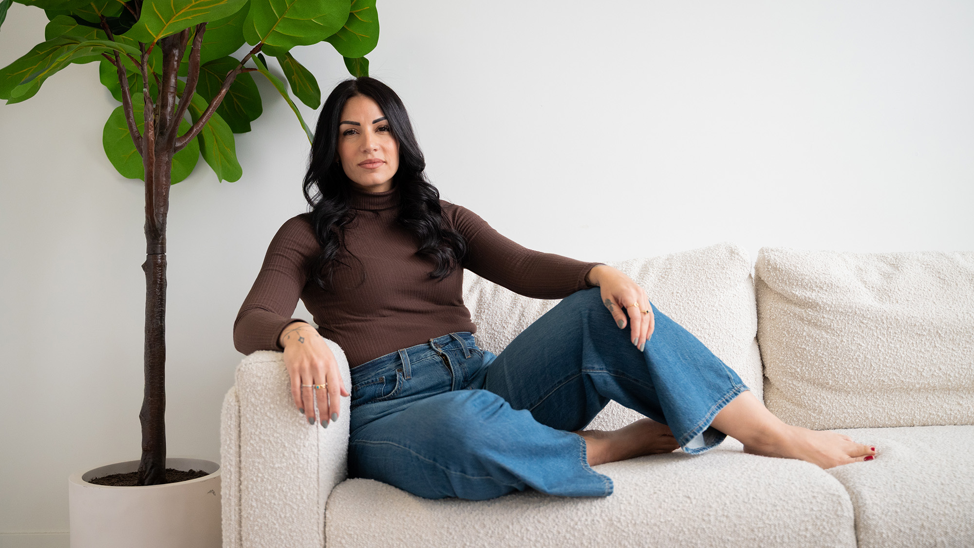 Tania Khazaal lounging on a plush white couch, wearing a brown knit turtleneck and jeans. She is barefoot and has long black hair. There is a large fake potted rubber tree plant to the left and slightly behind her.